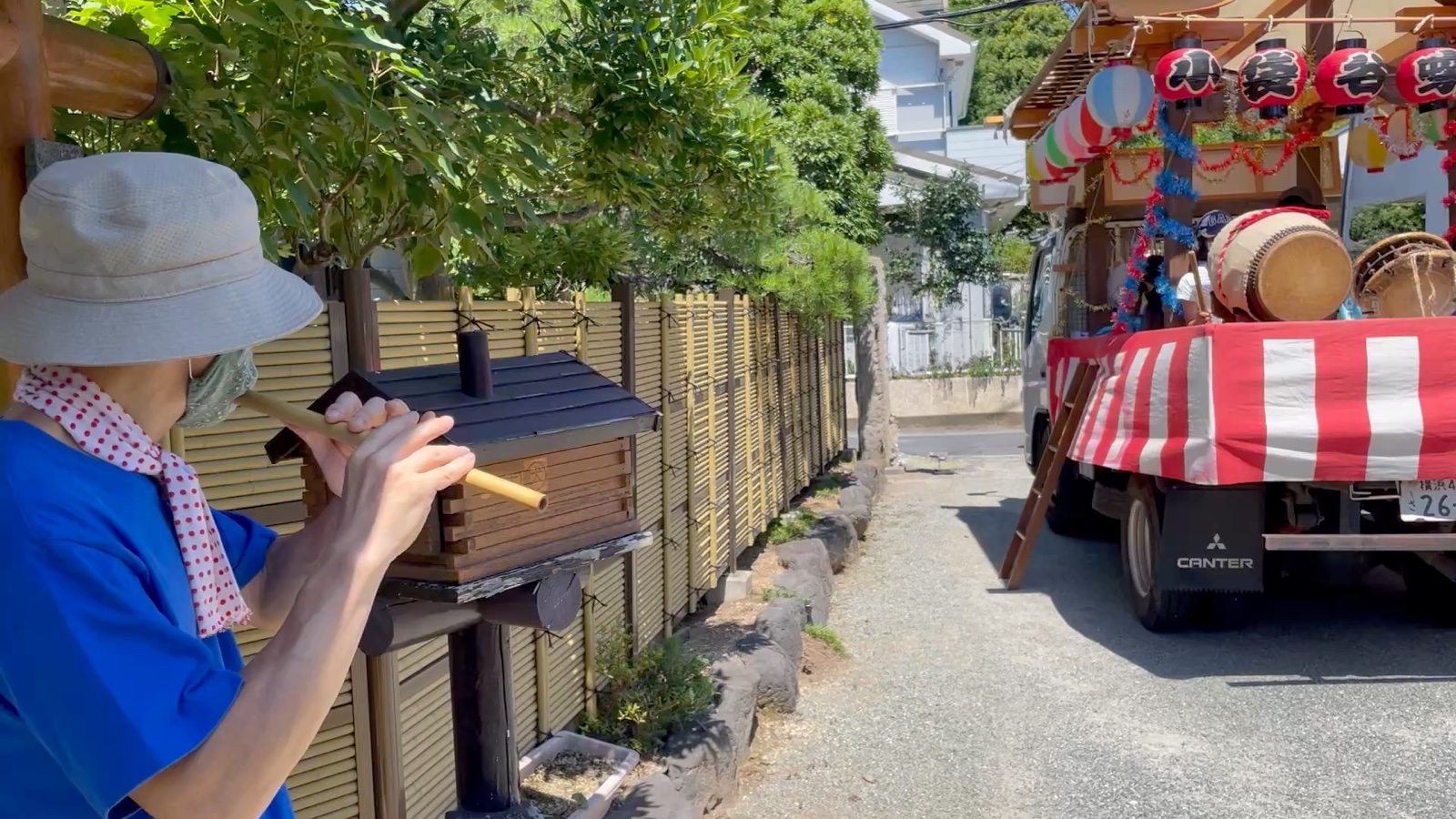 地域のお囃子の現場。小さな祠の前で篠笛を吹く人と、奥には屋台のトラック
