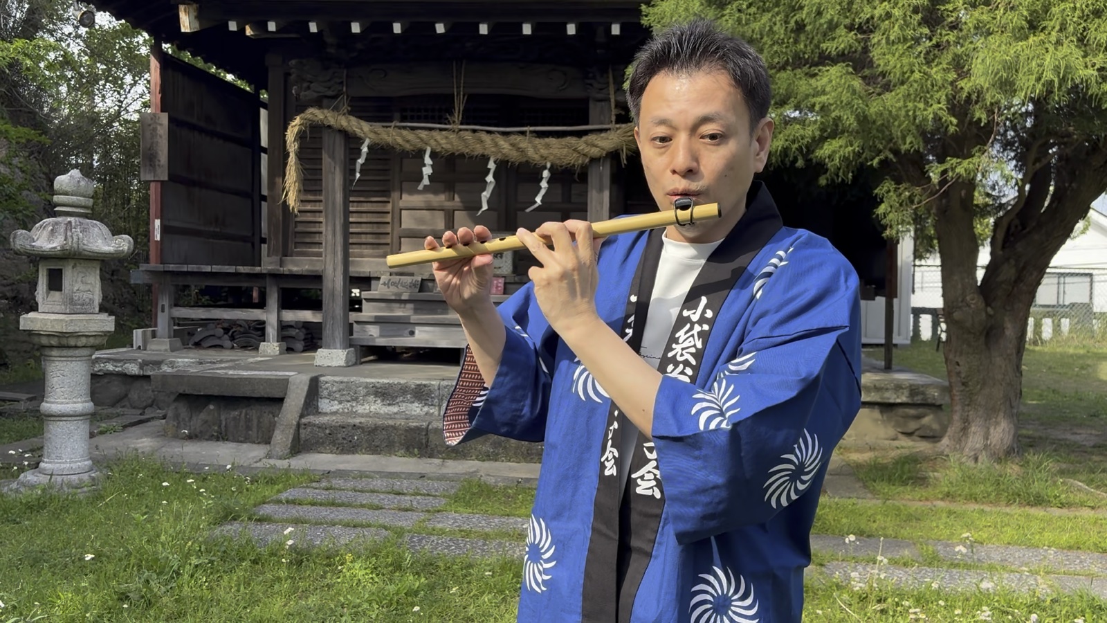法被姿で篠笛を吹く大谷さん。吹場鳴蔵で音が出せた瞬間、地域の祠の前にて