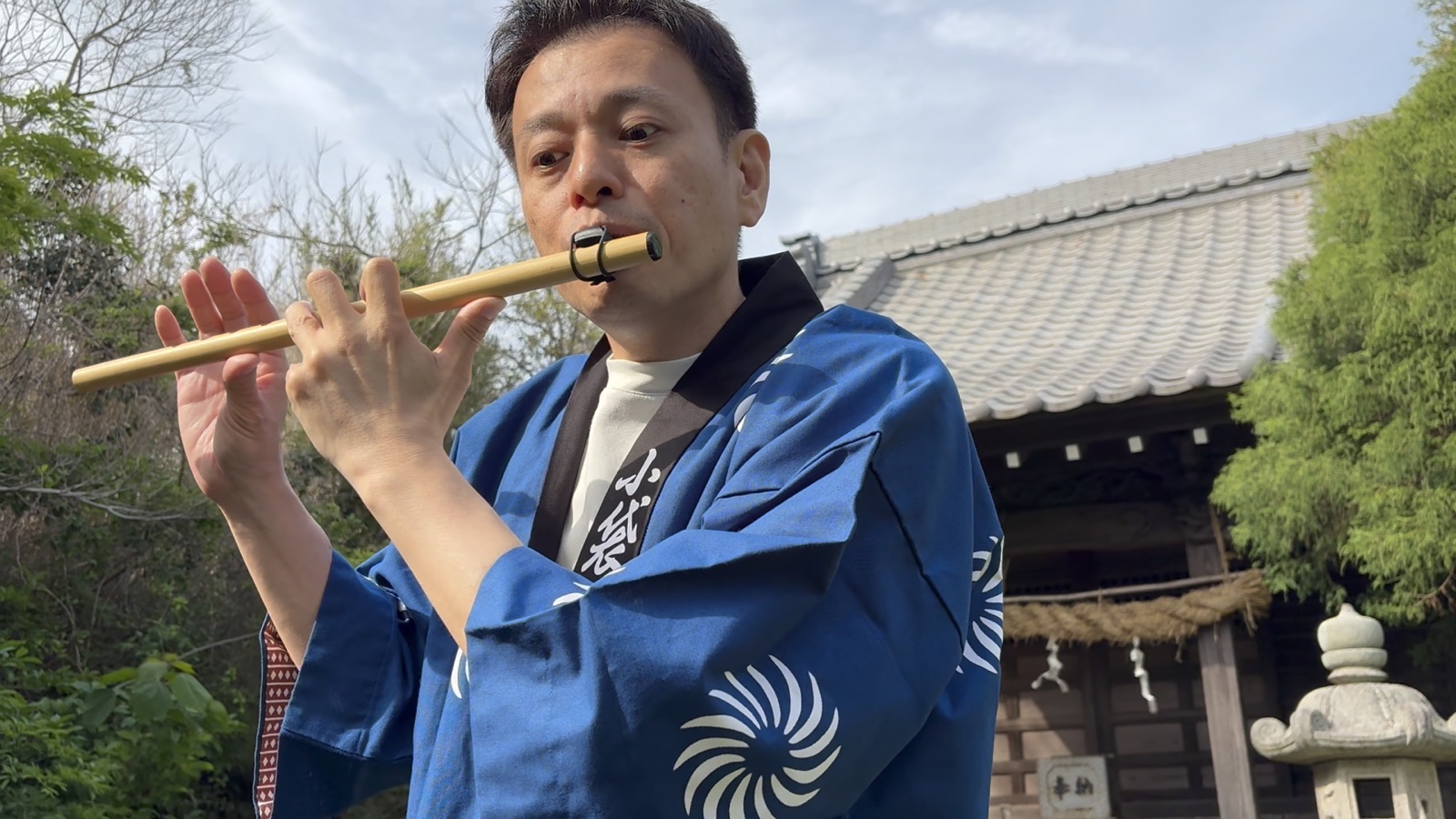 法被姿で篠笛を吹く大谷さん、地域の祠を背景に