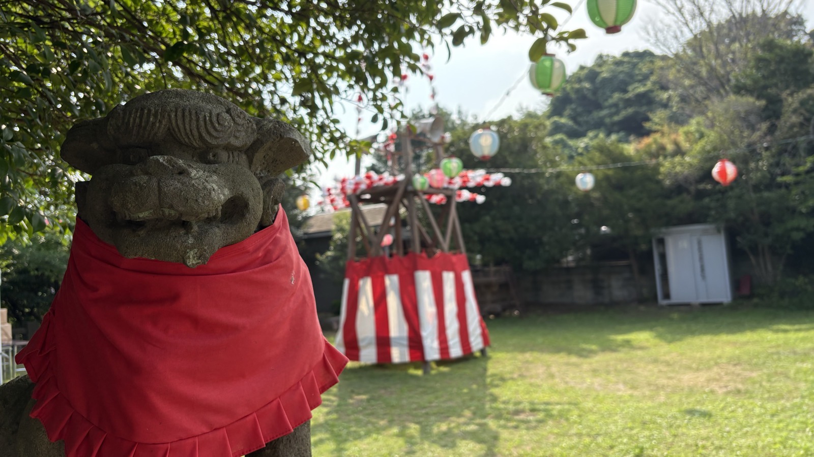 祭りの朝の境内。赤い前掛けをした狛犬の向こうに、紅白幕を巻いた櫓と、木々に渡された色とりどりの提灯が並ぶ